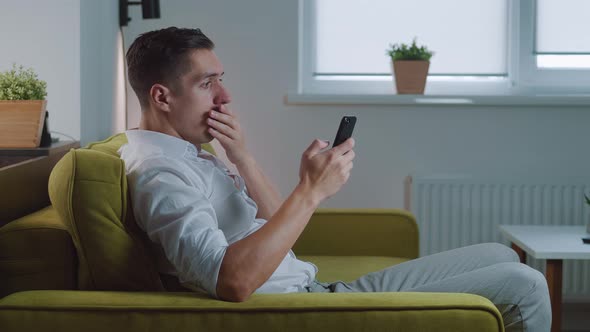 Shocked Man Reacting Message on Smartphone Covers His Mouth in Fright Sitting on Sofa at Home Living alt