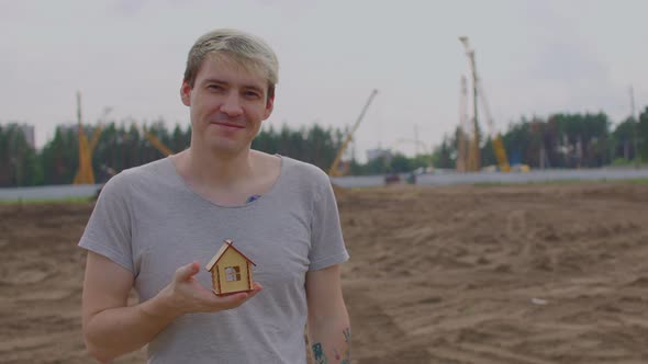 Young Man with Small Wooden House in Hand Stands on Construction Site alt