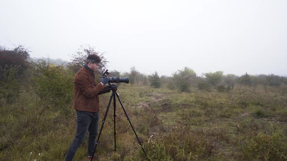 Wildlife photographer using DSLR camera on a tripod in a misty field. alt