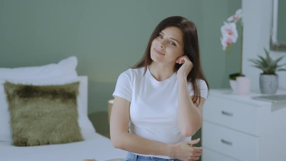 Pretty millennial brunette woman looking at the camera and smiling while sitting on bed at home alt
