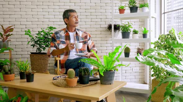An old man sitting in a living room decorated with flower pots. alt