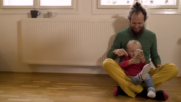 Father and Infant son sitting on floor using cell phone at living room. alt
