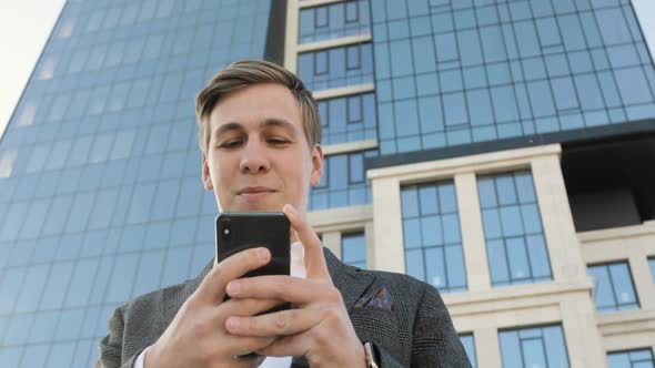 Young Businessman Is Typing on Smartphone Near Office Building in Downtown.