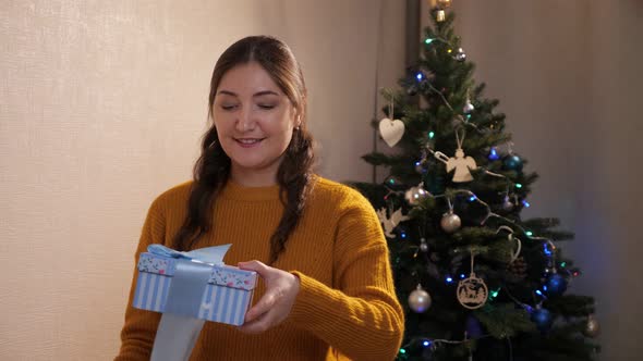 Woman in a Mustard Sweater Unties a Ribbon From a Gift Box Against Background of a Decorated Tree alt
