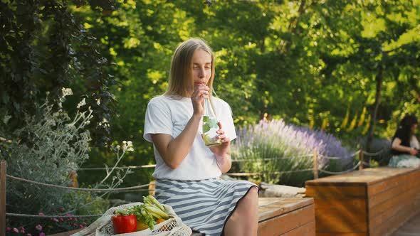 Woman with Vegetables and Greens in String Bag is Sitting on Bench of City Park alt