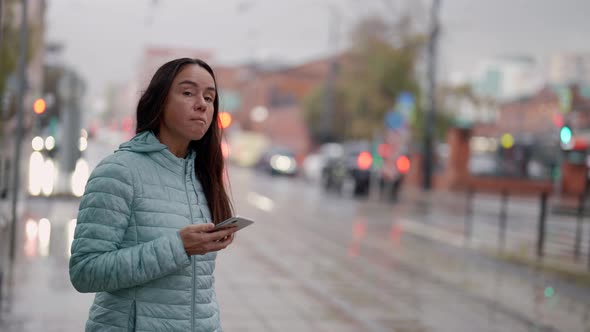 a Brunette in a Blue Jacket with Long Hair and with a Phone in Hand Stands on a Wet Street on a alt