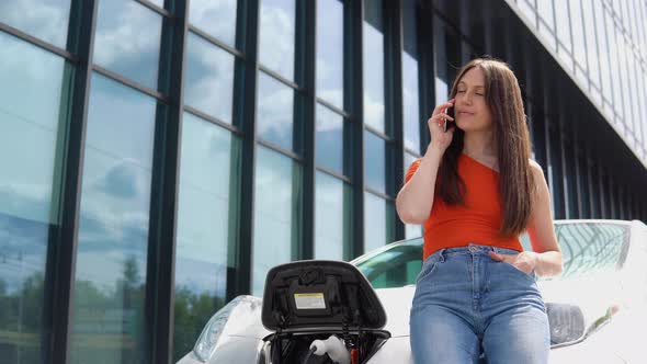 Attractive Woman Talking on Phone While Charging Electric Car Near the Shopping Center alt