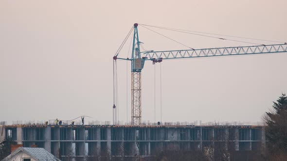 Dark Silhouette of Tower Crane and Small Silhouettes of Workers at High Residential Apartment alt