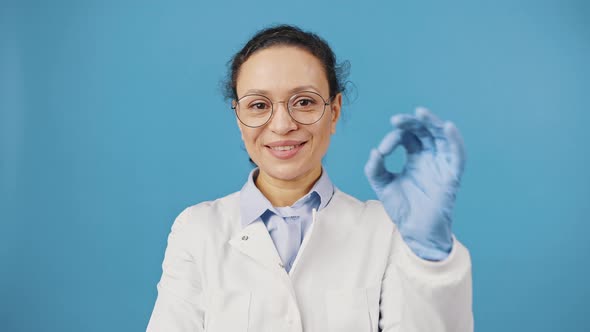 Positive Woman Doctor in Glasses Showing OK Gesture and Smiling to Camera Blue Studio Background alt