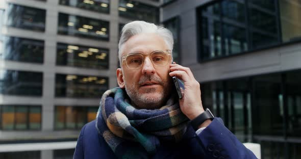 A Grayhaired Man Uses a Mobile Phone Standing in Business Center of the City alt