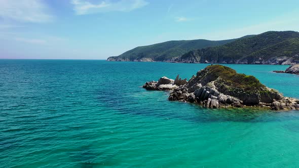 Small rocky island surrounded by crystal clear blue water with mountain and forest in the background alt