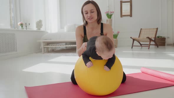 Cute Infant Boy Doing Exercises Lying on Fitness Ball While Mother Holding Her Legs alt