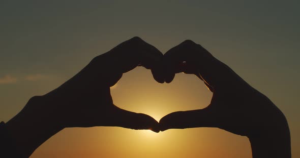 Close Up Female Hands Holding Heart Sign Silhouetted on Orange Sunset Sky Background. Faceless Woman