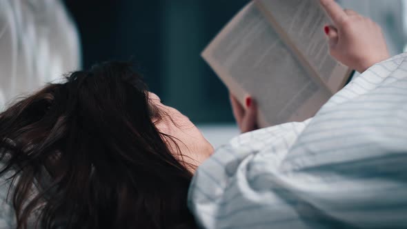 Brunette Woman Covered with a Blanket Lies on the Bed and Reads a Book at Night an Evening Alone