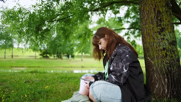 Brunette Woman Sits in the Park Under a Tree and Uses the Phone
