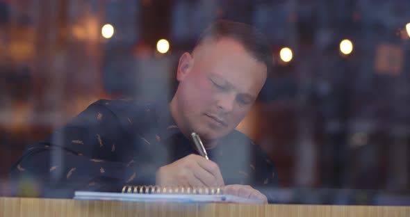 A Man Sits In A Cozy Cafe And Thinking About The Text And Writes It In A Notebook