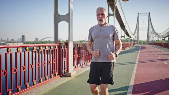 Elderly Man Jogs on Pedestrian Bridge Stopping to Rest alt