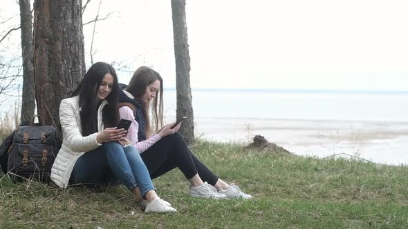 Two Caucasian female tourists sit cliff near tree and use their phones alt