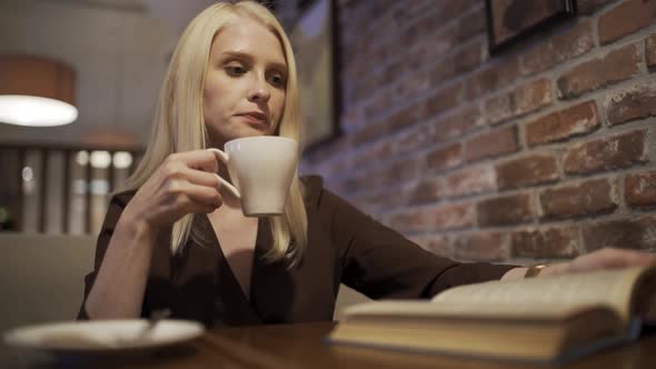 Beautiful Blonde Woman Enthusiastically Reads a Book and Drinks Aromatic Coffee