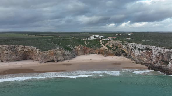 Wide aerial shot of surfers waiting in the waters of a beautiful bay surrounded by cliffs alt