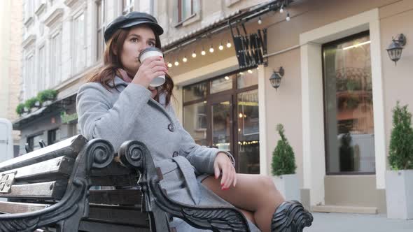 Young Beautiful Girl Drinking Coffee On The Street Sitting On A Bench.