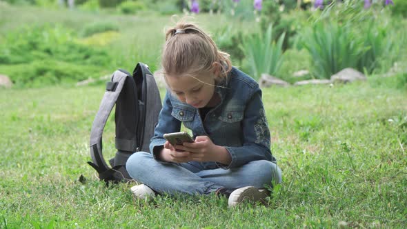 A pensive girl of nine years old, a schoolgirl, sits on the grass in the park.