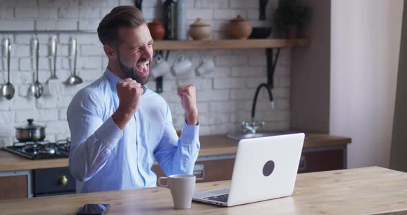 Young Business Man Sits at Table with a Laptop Working in Home Office Kitchen Background alt