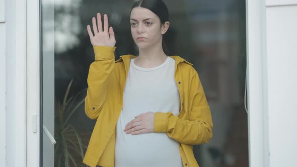 Young Sad Pregnant Woman Walking to Glass Door Touching Window Sighing Looking Out alt