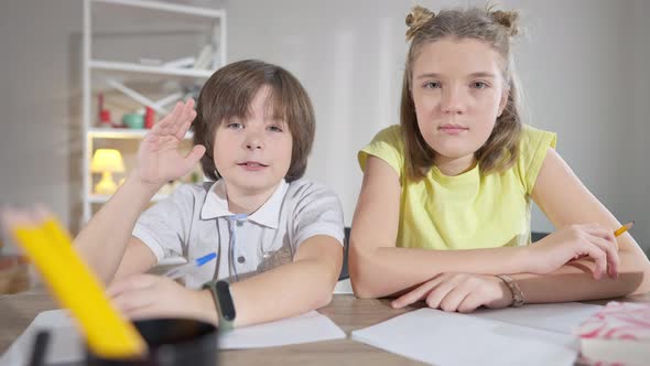 Brunette Caucasian Boy Raising Hand and Talking As Girl Sitting at Desk Looking at Camera alt