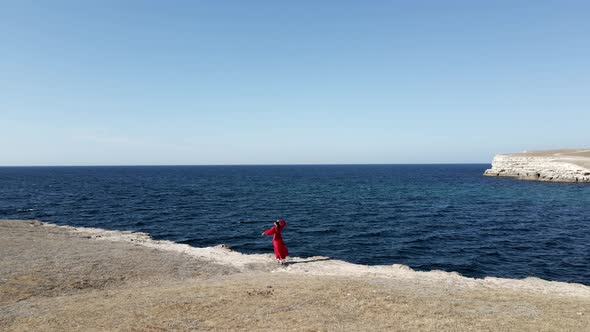 Brunette Woman in a Red Long Dress Stands on the Edge of a Cliff By the Sea alt