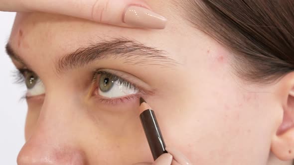 Professional Stylist Makeup Artist with Brown Pencil Draws Arrows in Eye Makeup to Young Beautiful alt