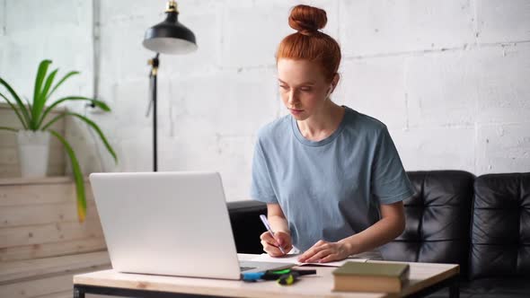 Busy Pretty Redhead Young Woman Student Is Taking Notes in Workbook By Looking at Laptop Screen. alt