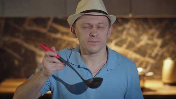 A Man in a Hat with a Ladle Stands in Front of the Kitchen Tasting Food From the Ladle alt