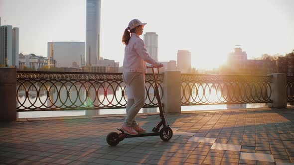 Attractive Woman Dressed in Pink Jacket Rides an Electric Scooter on Promenade alt