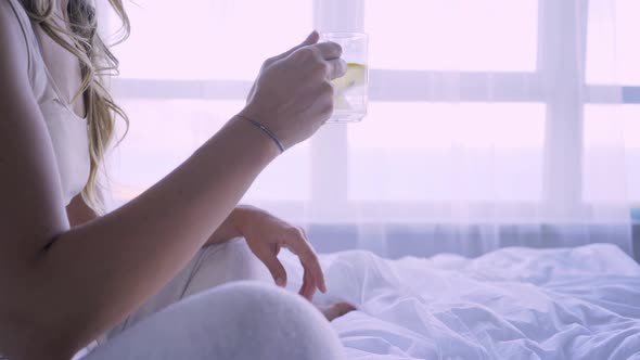 Young Woman with Curly Hair Hand Lifts Glass of Water on Bed