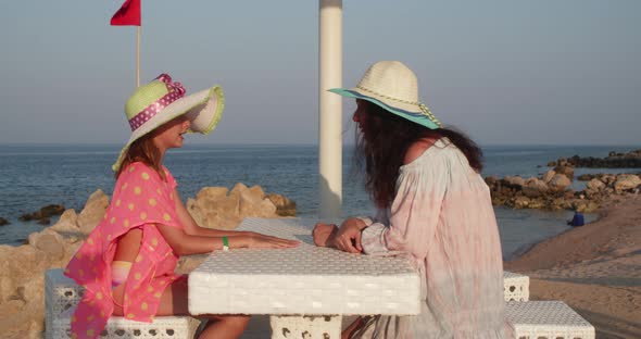 Woman is Talking to Her Teen Daughter Sitting at Table on Resort Sea Beach