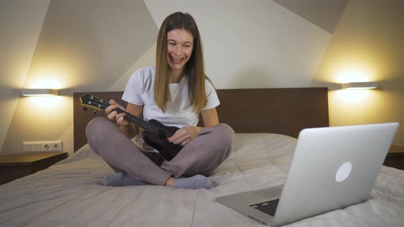 Joyful Young Woman Playing The Ukulele In House On Bed And Using Laptop in Quarantine. Distance