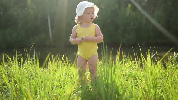 Little Funny Cute Blonde Girl Child Toddler in Yellow Bodysuit and White Hat Walking in Field with
