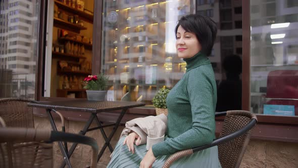 Pretty Woman in Green Sits in a Cafe on the Street Looks at the Camera and Smiles
