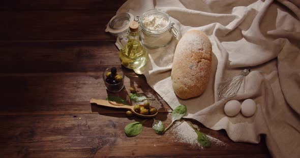 Wholemeal Bread On A Wooden Table, A Bottle Of Oil, Olives, Flour And Eggs On A Linen Tablecloth