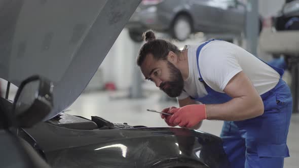 Adult Mechanic Standing Near the Car with an Open Hood and Verifies Condition of Engine alt