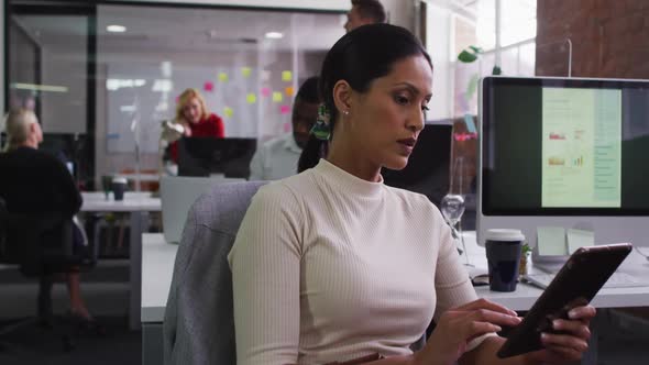 Mixed race businesswoman sitting at desk in open space using tablet smiling alt