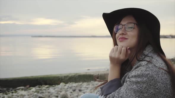 Young Pretty Smiling Woman in Black Hat and Glasses Sitting Near Sea at Sunset alt