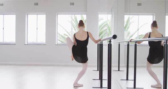 Caucasian female ballet dancer stretching up by the mirror in a bright studio alt