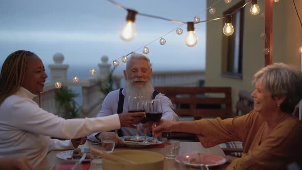 Happy multiracial seniors toasting with red wine glasses together on house patio dinner alt