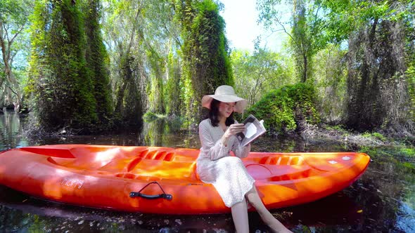 Asian woman, a tourist, reading a book on a boat or kayak with trees in Rayong Botanical Garden alt