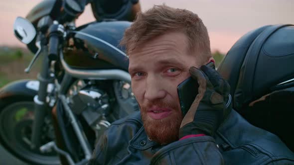 A Middleaged Male Biker Sits By a Motorcycle and Talks on His Mobile Phone