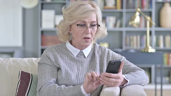 Old Woman Celebrating Success on Smartphone on Sofa alt