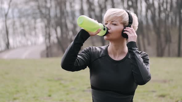 Athletic Woman in a Mask Is Listening To Music in Earphones and Drinking Water alt