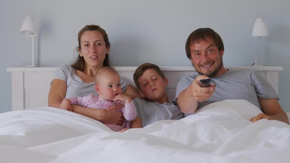 Happy Young European Family with Two Kids Gather on Couch to Watch TV in the Badroom Smiling alt